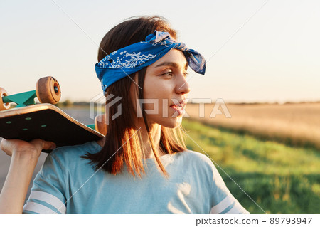 Side view portrait of pensive beautiful female wearing blue casual t shirt and stylish hair band, looking away with thoughtful look, holding skateboard over shoulders. Side view portrait of pensive beautiful female wearing blue casual t shirt and stylish hair band, looking away with thoughtful look, holding skateboard over shoulders. 89793947