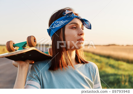 Pretty dark haired female wearing t shirt and hair band holding skateboard on her shoulder and looking away with pensive facial expression, resting after skateboarding. 89793948