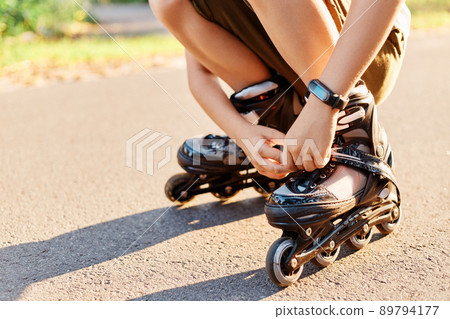 Close up portrait of unknown child squats on road and fixing laces on roller blades before skating, faceless kid having fun outdoor, rollerblading, playing alone. 89794177