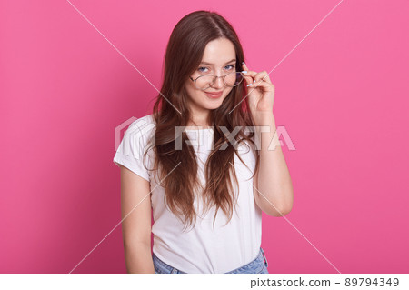 Indoor shot of attractive young woman with long hair keeps hand on rim of spectacles, wearing casual outfit, model posing against pink studio wall, looks smiling directly at camera. People concept. Indoor shot of attractive young woman with long hair keeps hand on rim of spectacles, wearing casual outfit, model posing against pink studio wall, looks smiling directly at camera. People concept. 89794349