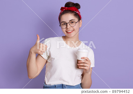 Happy young woman showing thumb up and holding take away coffee, looking smiling directly at camera, wearing casual attire and red hairband, being in good mood. 89794452