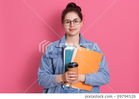Young attractive student girl looking directly at camera with smile, posing isolated over rose background, wearing denim jacket and spectacles, holding paper folder and take away coffee. 89794526