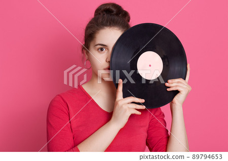 Brunette lady with knot dresses red casual shirt looking directly at camera with serious facial expression, covering half of face with retro music plate. Brunette lady with knot dresses red casual shirt looking directly at camera with serious facial expression, covering half of face with retro music plate. 89794653