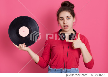 Astonish young charming girl holding music plate in hand and pointing at it with forefinger, keeps mouth opened, sees vintage disc for first time, being surprised. Astonish young charming girl holding music plate in hand and pointing at it with forefinger, keeps mouth opened, sees vintage disc for first time, being surprised. 89794656