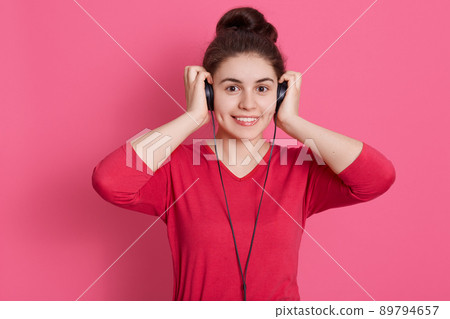 Teenage girl with charming smile standing against pink background with head phones, touching her headset with palms, looks happy, enjoying to listen to favorite music. Teenage girl with charming smile standing against pink background with head phones, touching her headset with palms, looks happy, enjoying to listen to favorite music. 89794657