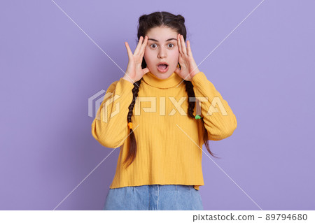 Stunned dark haired woman with two pigtails, keeping hands near face, afraids of something astonishing, dresses in fashionable yellow shirt, posing isolated over lilac background. 89794680