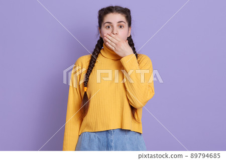Young beautiful brunette woman wearing casual yellow sweater, posing over lilac background, being shocked, female covering mouth with hands for mistake. Young beautiful brunette woman wearing casual yellow sweater, posing over lilac background, being shocked, female covering mouth with hands for mistake. 89794685