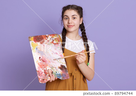 Indoor shot of professional female artist smiling with paint brush and palette. Woman with two pigtails painting in her workshop, looks at camera, stands against lilac wall. Indoor shot of professional female artist smiling with paint brush and palette. Woman with two pigtails painting in her workshop, looks at camera, stands against lilac wall. 89794744
