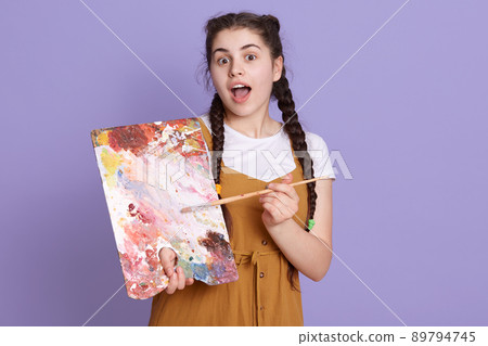 Young brunette artist woman with pigtails holding painter brush and palette over lilac background, posing with surprise face, standing with opened mouth. Young brunette artist woman with pigtails holding painter brush and palette over lilac background, posing with surprise face, standing with opened mouth. 89794745