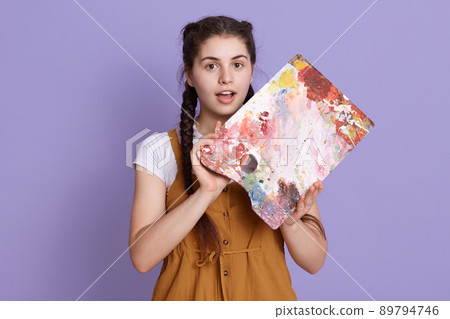 Astonished woman with painting palette in hands, keeps mouth opened, looks surprised, standing in casual clothing against lilac wall, expressing astonish. Astonished woman with painting palette in hands, keeps mouth opened, looks surprised, standing in casual clothing against lilac wall, expressing astonish. 89794746