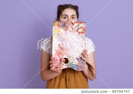 Portrait of attractive young female artist hiding behind color palette, looking at camera, adorable female wearing casual attire, posing isolated over lilac background. Portrait of attractive young female artist hiding behind color palette, looking at camera, adorable female wearing casual attire, posing isolated over lilac background. 89794747