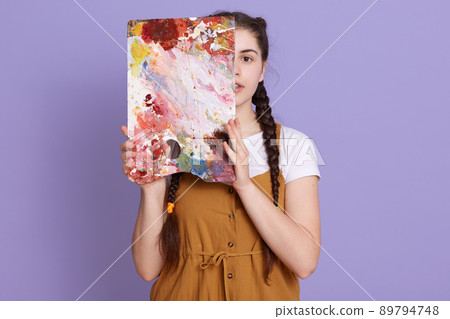 Young woman covering half of her face with easel, looks at camera, wearing casual clothing and standing isolated over lilac background, artist posing with her picture. Young woman covering half of her face with easel, looks at camera, wearing casual clothing and standing isolated over lilac background, artist posing with her picture. 89794748