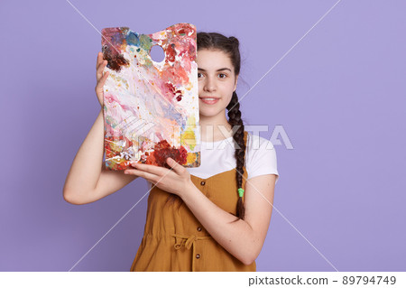 Picture of tender young lady holding picture in hands and covering half of face, wearing dress, standing isolated over lilac background, adorable artist showing her work. 89794749