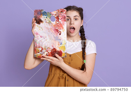 Indoor shot of creative young woman painting in her studio. Female artist in her workshop holding colors palette near her face, stands with widely open mouth, expresses shock, poses against lilac wall Indoor shot of creative young woman painting in her studio. Female artist in her workshop holding colors palette near her face, stands with widely open mouth, expresses shock, poses against lilac wall 89794750