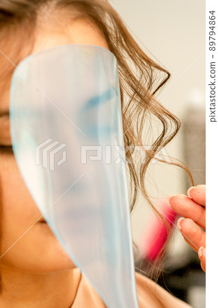 Professional hairdresser styling and fixing with hairspray long woman curly hair covering the face with a protective shield in a beauty salon 89794864