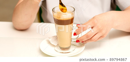 Woman with the latte. Glass mug of latte coffee on the white saucer with female hands holding teaspoon on the table in the cafeteria, close up 89794867