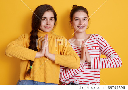 Two young females wearing casual shirts posing with palms together and looking smiling directly at camera, ladies practicing yoga, standing against yellow wall. Two young females wearing casual shirts posing with palms together and looking smiling directly at camera, ladies practicing yoga, standing against yellow wall. 89794896