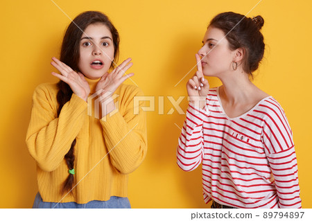 Attractive female wearing striped shirt keeps finger near lips while telling secret to her friend, girl in yellow sweater being in shock, keeps mouth opened, expressing astonish. Attractive female wearing striped shirt keeps finger near lips while telling secret to her friend, girl in yellow sweater being in shock, keeps mouth opened, expressing astonish. 89794897
