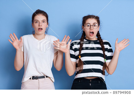 European dark haired girls posing with hands up, widely opened mouth, having shocked facial expressions, express fear, wearing casual t shirts, posing against blue wall. 89794908