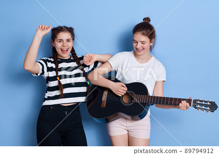 Two friends singing and dancing isolated over blue background, lady with knot playing guitar, winsome girl in striped t shirt and pigtails raising hands up. 89794912