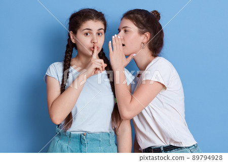 Two friends gossiping isolated over blue background, girl with knot whispering secret and keeps palm near mouth, girl with pigtails listening to her friend and keeping finger near lips. 89794928