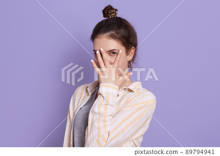 Attractive lady covering her face with fingers, woman with dark hair and bun, female posing isolated over lilac background, girl wearing white casual shirt. Attractive lady covering her face with fingers, woman with dark hair and bun, female posing isolated over lilac background, girl wearing white casual shirt. 89794941