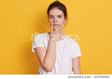 Serious young woman wearing white t shirt looking at camera with confident expression, looks thoughtful, thinks about solving problem or finds answer question, stands against yellow wall. Serious young woman wearing white t shirt looking at camera with confident expression, looks thoughtful, thinks about solving problem or finds answer question, stands against yellow wall. 89794980