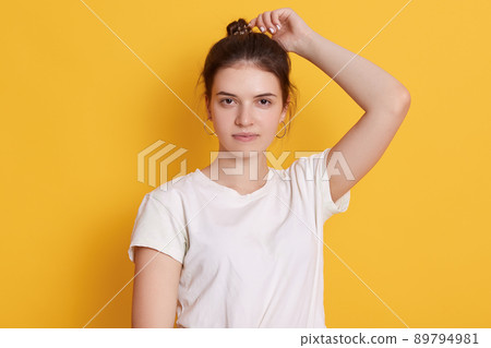 Confident female being photographed against yellow wall, keeping arm up, looking directly at camera, wearing casual white t shirt, having dark hair and bun. Confident female being photographed against yellow wall, keeping arm up, looking directly at camera, wearing casual white t shirt, having dark hair and bun. 89794981
