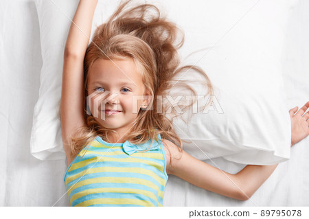 Above view of attractive light haired small child, has blue eyes, stretches in bed, lies on white pillow, dressed in colourful striped pyjamas, enjoys recreation time in bedroom. Children and rest Above view of attractive light haired small child, has blue eyes, stretches in bed, lies on white pillow, dressed in colourful striped pyjamas, enjoys recreation time in bedroom. Children and rest 89795078