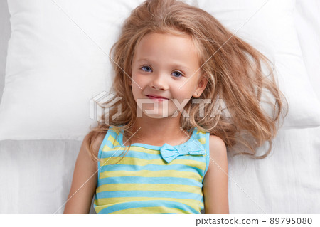 Photo of pleasant looking small female kid with blue eyes, dressed in nightwear, has satisfied expression, lies on white bedclothes, enjoys good rest, rejoices starting new day. Bed time and children Photo of pleasant looking small female kid with blue eyes, dressed in nightwear, has satisfied expression, lies on white bedclothes, enjoys good rest, rejoices starting new day. Bed time and children 89795080