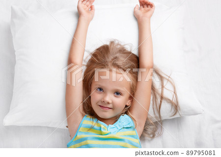 Top view of pretty child has long hair, stretches in bed, lies on white pillow, has good mood after rest in bed, awakes early in morning, wants to start new day, looks directly at camera. Childhood Top view of pretty child has long hair, stretches in bed, lies on white pillow, has good mood after rest in bed, awakes early in morning, wants to start new day, looks directly at camera. Childhood 89795081