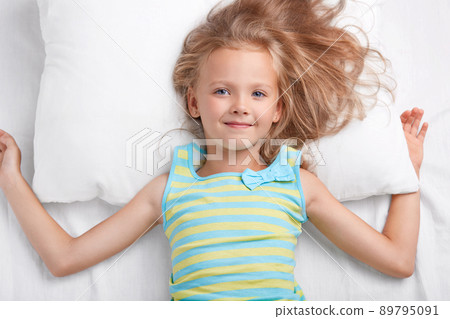 Children, bedding and awakening concept. Pleased small European girl with charming tender smile, long light hair, dressed in pyjamass, lies on white pillow and bedclothes, has appealing appearance. Children, bedding and awakening concept. Pleased small European girl with charming tender smile, long light hair, dressed in pyjamass, lies on white pillow and bedclothes, has appealing appearance. 89795091