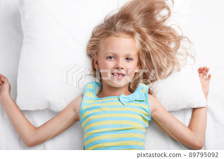 Photo of small cute female child has attractive look, lies in comfortable bed on pillow, dressed in nightwear, enjoys good morning, smiles positively at camera. Children and awakening concept Photo of small cute female child has attractive look, lies in comfortable bed on pillow, dressed in nightwear, enjoys good morning, smiles positively at camera. Children and awakening concept 89795092