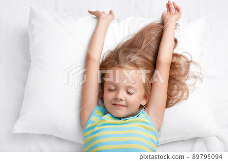 Beautiful small adorable kid stretches in bed, dressed in casual pyjamass, lies on white comfortable pillow, keeps eyes shut, being in bedroom, has healthy sleep. Good morning and happiness concept 89795094