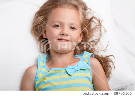 Close up shot of charming small kid with healthy skin, light hair, blue appealing eyes, healthy soft skinm wears pyjamass, lies on white bedclothes in bedroom, awakes after having sleep. Awakening Close up shot of charming small kid with healthy skin, light hair, blue appealing eyes, healthy soft skinm wears pyjamass, lies on white bedclothes in bedroom, awakes after having sleep. Awakening 89795106
