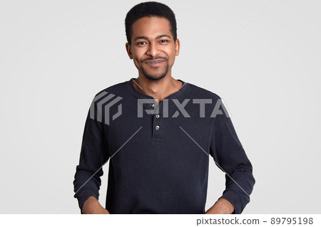 Photo of satisfied dark skinned guy with stubble, feels happy and pleased, being in good mood, wears casual clothes, models against white background. Afro American hipster smiles gently, stands indoor Photo of satisfied dark skinned guy with stubble, feels happy and pleased, being in good mood, wears casual clothes, models against white background. Afro American hipster smiles gently, stands indoor 89795198
