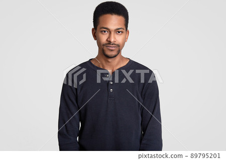 Waist up shot of black man with short curly hair, little beard, looks seriously at camera, thinks about something with contemplative facial expression, dressed in casual outfit, isolated on white wall Waist up shot of black man with short curly hair, little beard, looks seriously at camera, thinks about something with contemplative facial expression, dressed in casual outfit, isolated on white wall 89795201