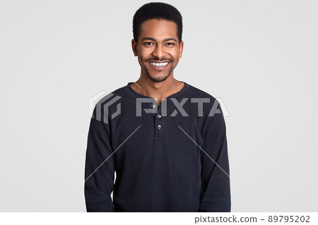 Smiling Afro American man has toothy smile, shows white teeth, being in high spirit, rejoices pleasant moments in life, wears casual black jumper, models against white background. Happiness concept Smiling Afro American man has toothy smile, shows white teeth, being in high spirit, rejoices pleasant moments in life, wears casual black jumper, models against white background. Happiness concept 89795202