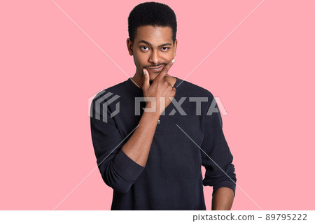 Attractive black man holds chin, looks with puzzlement, raises eyebrow, tries to find necessary solution of problem, wears casual jumper, isolated over pink backgrund. People, thoughts, decision 89795222