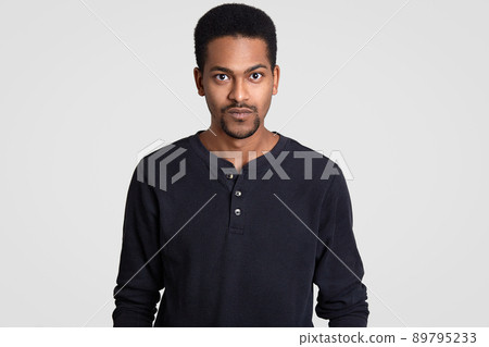 Indoor shot of serious self confident dark skinned young man with curly short hair, looks with stern expression, models indoor against white background. People, ethnicity and lifestyle concept Indoor shot of serious self confident dark skinned young man with curly short hair, looks with stern expression, models indoor against white background. People, ethnicity and lifestyle concept 89795233