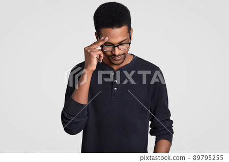 Stressful tired overworked male worker has dark skin, keeps hand on head, looks down, wears transparent glasses and casual jumper, tries to gather with thoughts, isolated over white background Stressful tired overworked male worker has dark skin, keeps hand on head, looks down, wears transparent glasses and casual jumper, tries to gather with thoughts, isolated over white background 89795255