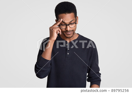 Depressed African American male keeps hand on forehead, recollects information in mind, feels displeased, focused down, being tired, suffers from headache, dressed in casual outfit, poses indoor 89795256