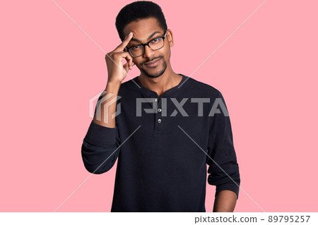 Dark skinned handsome male looks thoughtful. Man wears glasses and black sweater. Young guy with beard keeps fore finger on forehead. Model poses in photo studio isolated over rose background. Dark skinned handsome male looks thoughtful. Man wears glasses and black sweater. Young guy with beard keeps fore finger on forehead. Model poses in photo studio isolated over rose background. 89795257