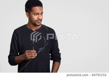 Studio shot of black male director focused thoughtfully aside, holds spectacles in hands, being deep in thoughts, wars casual jumper, isolated over white background with free space for text. 89795258