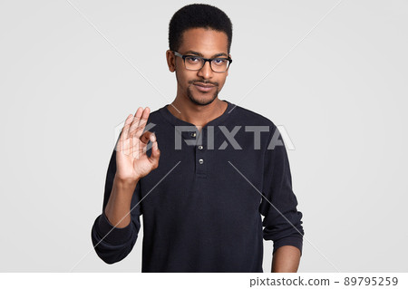 Waist up shot of confident dark skinned man shows approval gesture, makes okay sign, dressed in casual outfit, feels self confident, wears transparent glasses, isolated over white wall. Body language Waist up shot of confident dark skinned man shows approval gesture, makes okay sign, dressed in casual outfit, feels self confident, wears transparent glasses, isolated over white wall. Body language 89795259