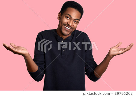 Young dark skinned male in confusion, stands smiling isolated over pink background. Handsome guy wears casual black shirt and makes helpless gesture. Model poses in photo studio. People concept. Young dark skinned male in confusion, stands smiling isolated over pink background. Handsome guy wears casual black shirt and makes helpless gesture. Model poses in photo studio. People concept. 89795262
