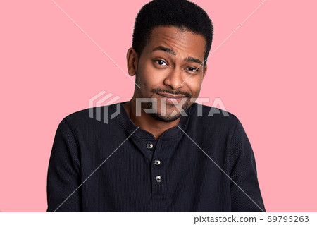 African man with beard and mustache crews his eyes and smiles, male looks shy. Model stands isolated over pink blackground, poses and looks at camera. Young guy wears black shirt. People concept. African man with beard and mustache crews his eyes and smiles, male looks shy. Model stands isolated over pink blackground, poses and looks at camera. Young guy wears black shirt. People concept. 89795263