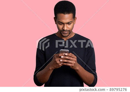 Young guy looks at the screen of smartphone with serious fecial expression. Black handsome male has important unpleasent correspondence. Boring pastime. Modern technology and Internet concept. 89795276