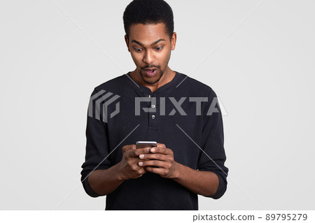 Surprised black man holds modern cellular, recieves unexpected text message, surfes web page, dressed in casual clothes, feels shocked, connected to wireless intenet, isolated over white background 89795279