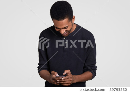 Overjoyed dark skinned man with Afro haircut, laughs happily, holds cell phone, reads funny story on cellular, types feedback, wears casual clothes, models over white background. Happiness concept Overjoyed dark skinned man with Afro haircut, laughs happily, holds cell phone, reads funny story on cellular, types feedback, wears casual clothes, models over white background. Happiness concept 89795280